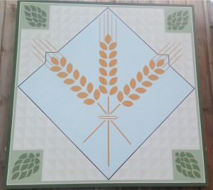 A square featuring three barley leaves intertwined. Behind the leaves is a blue square. at each corner there is a green hop bud
