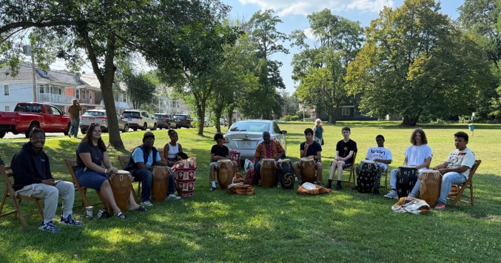 A group of teenagers sit with drums between their legs in a semi circle. Zorkie Nelson sits directly in the middle